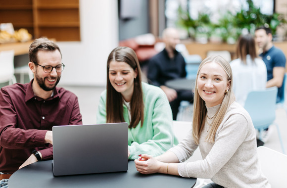 Drei Menschen schauen gemeinsam auf einen Laptop, eine Frau schaut direkt in die Kamera. Sie befinden sich in einem großen, hellen, freundlichen Atrium. Im Hintergrund besprechen sich noch weitere Menschen.